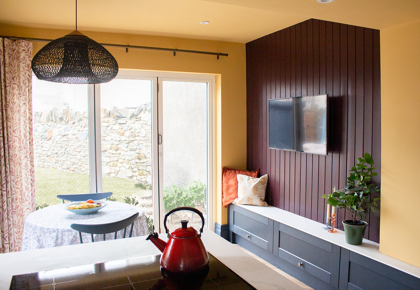 A kitchen with patterned curtains, a bench seat with cushions and round dinning table