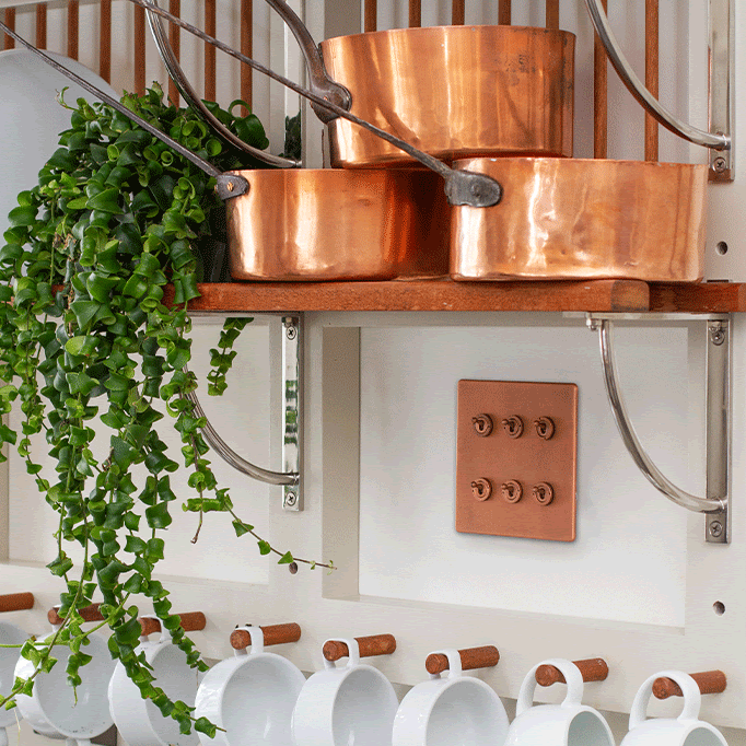 A photo of three copper pans on a shelf with matching copper light switches beneath the shelf.