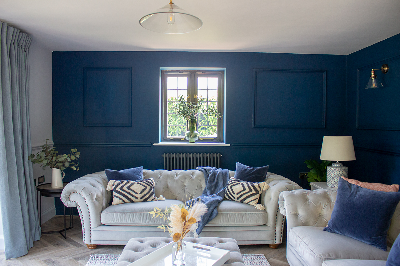 A photo of a living room with a dark blue wall with a column style radiator on it, behind a grey velvet sofa.