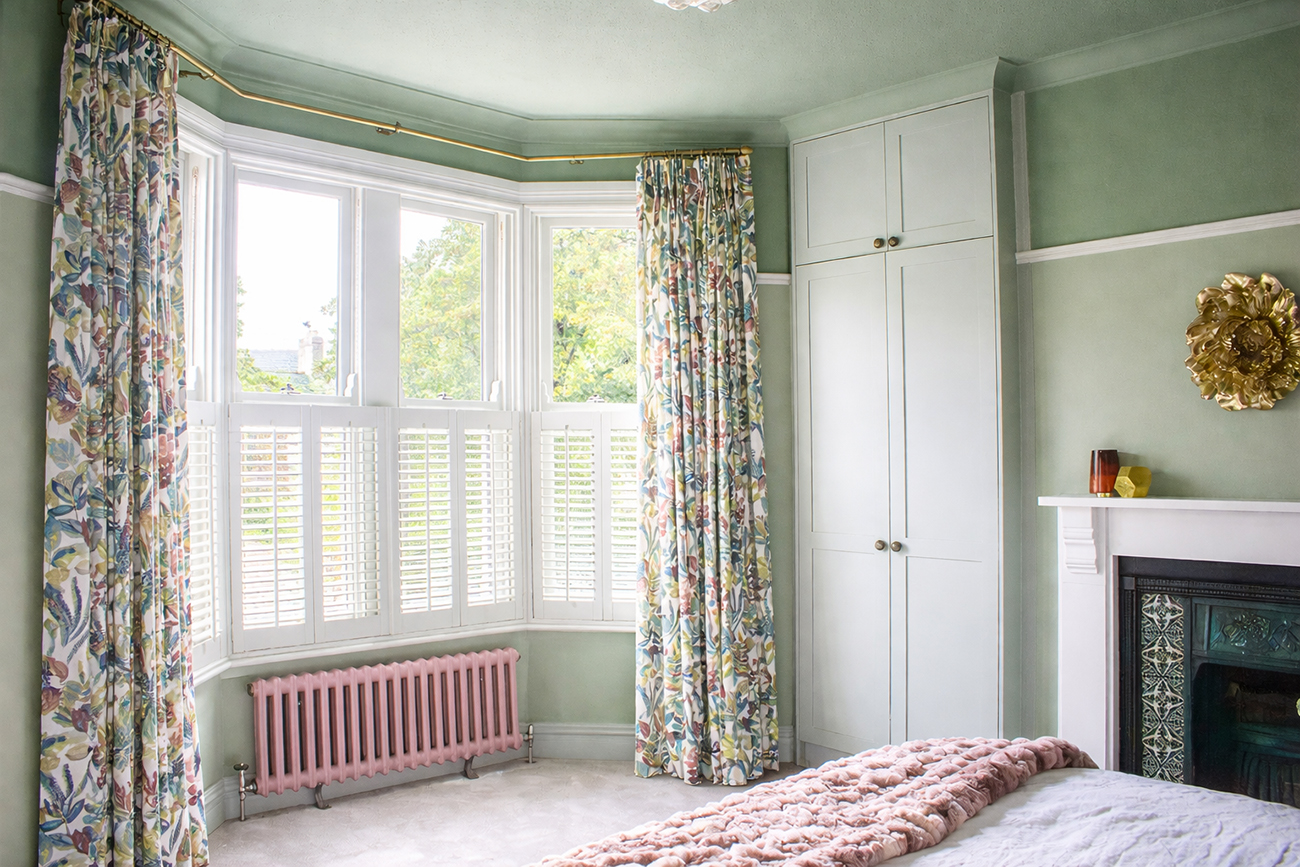 A photo of a bay window in a bedroom with pale green walls, a brass curtain pole and bold floral curtains.