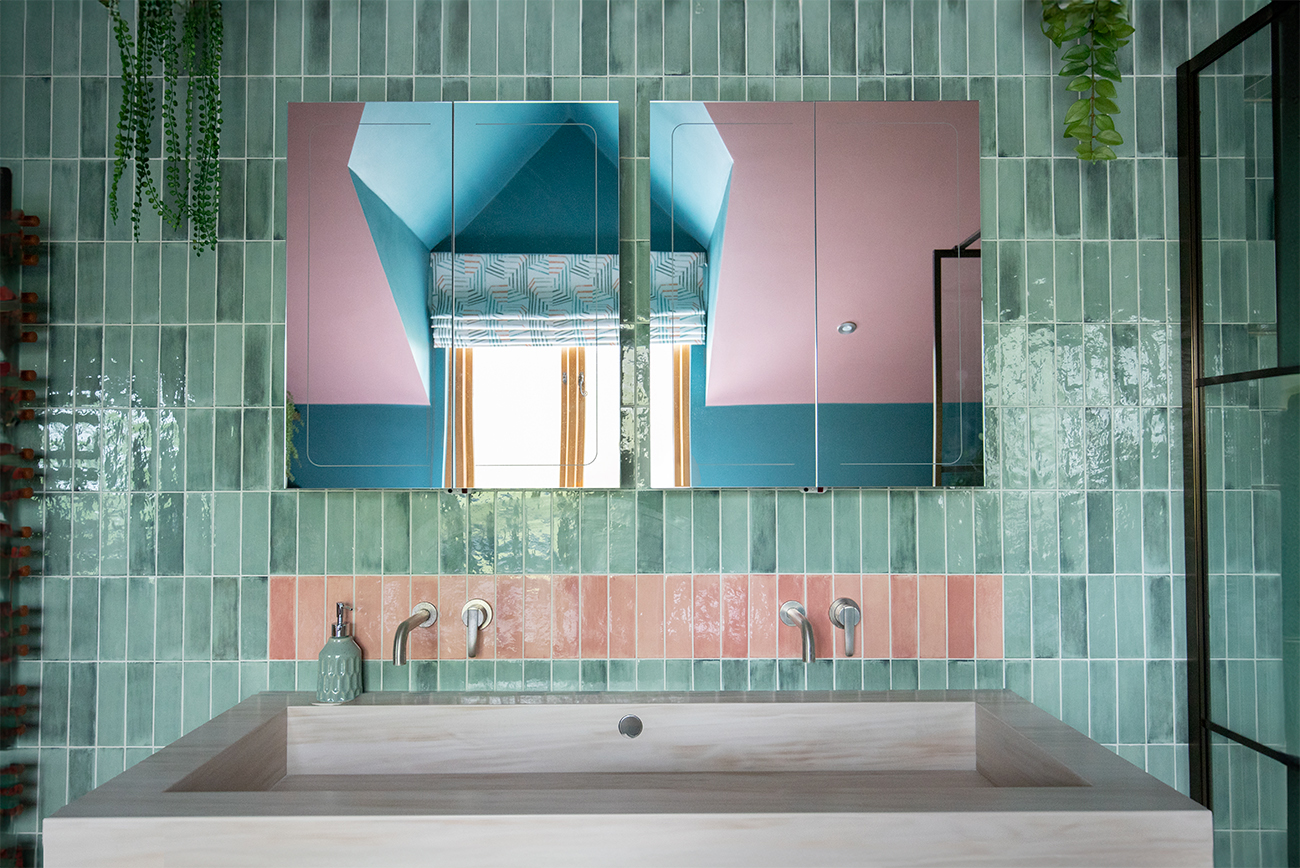 A photo of a bathroom with mid green tiles stacked vertically, with a row of pink tiles laid where the wall mounted taps are.