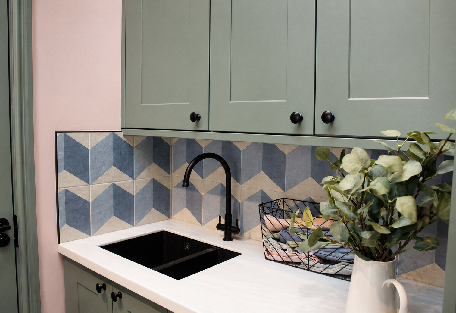  A photo of a utility room with green cabinets and pink walls, with a geometric blue tile and black tile trim.