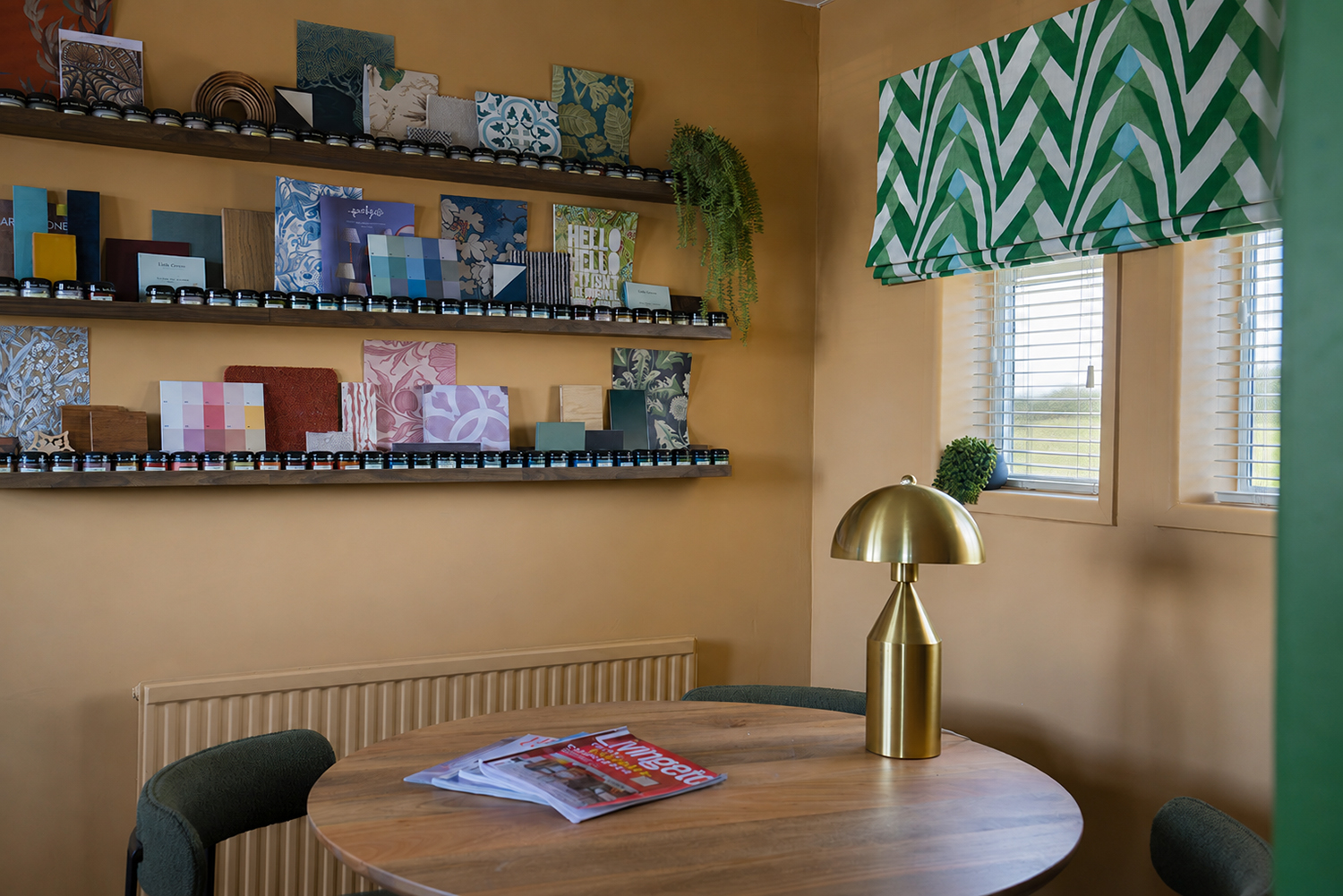 A photo of a small office with mustard yellow walls and a green blind, with a round walnut table and chairs.
