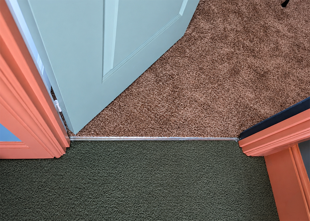 A photo looking down onto a green landing carpet and a blush pink office carpet, with a stain nickel door bar joining them.