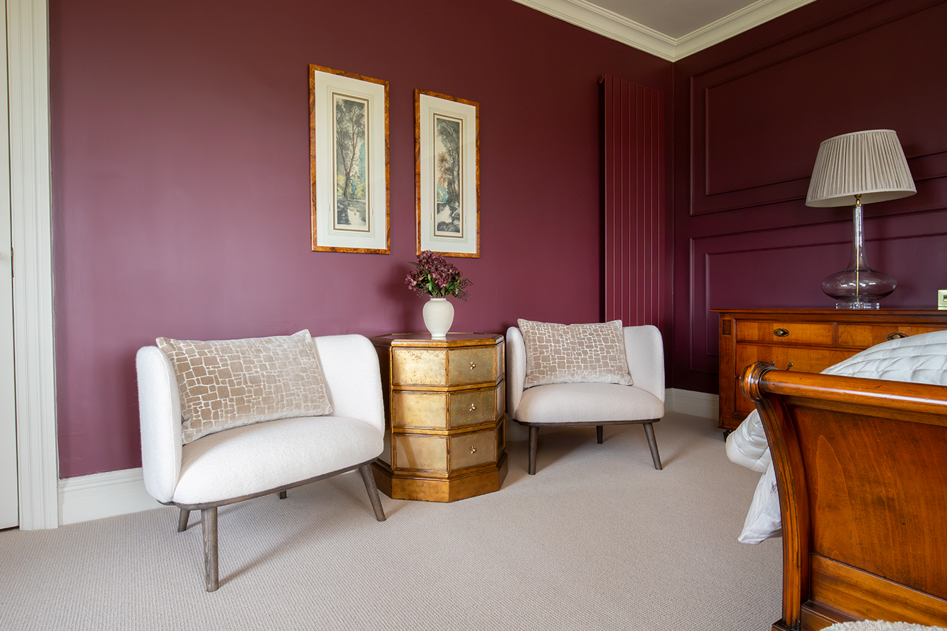 A photo of a bedroom with deep burgundy walls and cream armchairs, with a cream loop carpet.