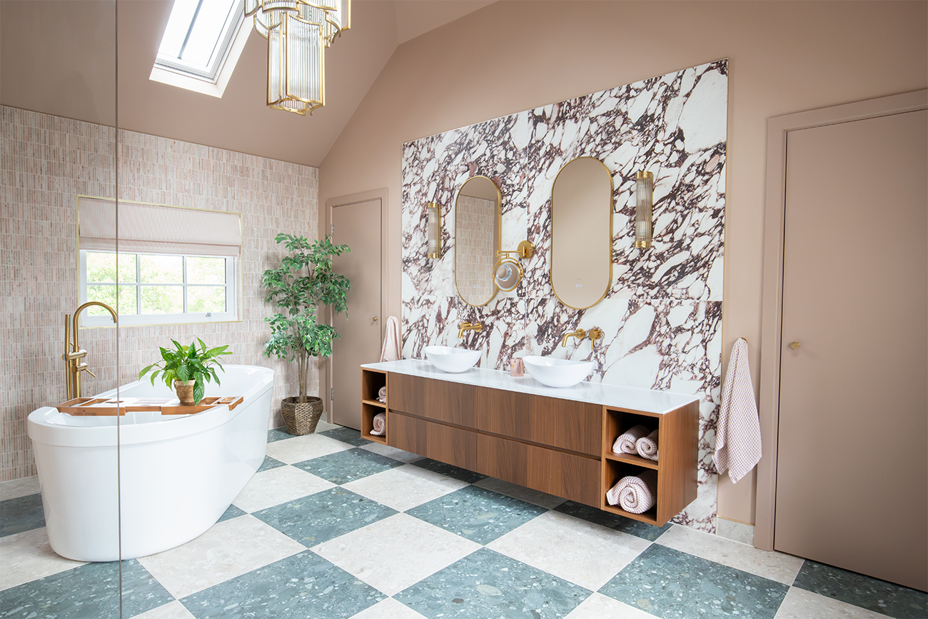 A photo of the en suite with warm mocha walls and dramatic burgundy veined tiles behind a walnut double vanity unti, and a frestanding bath to the left.