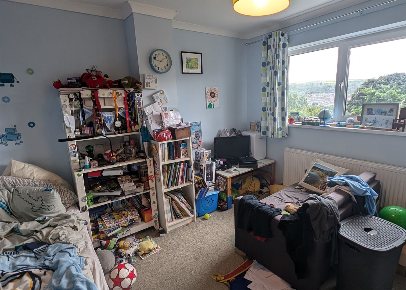 The pale blue bedroom before the redesign, with overfull bookshelves and a small gaming set up in the corner.