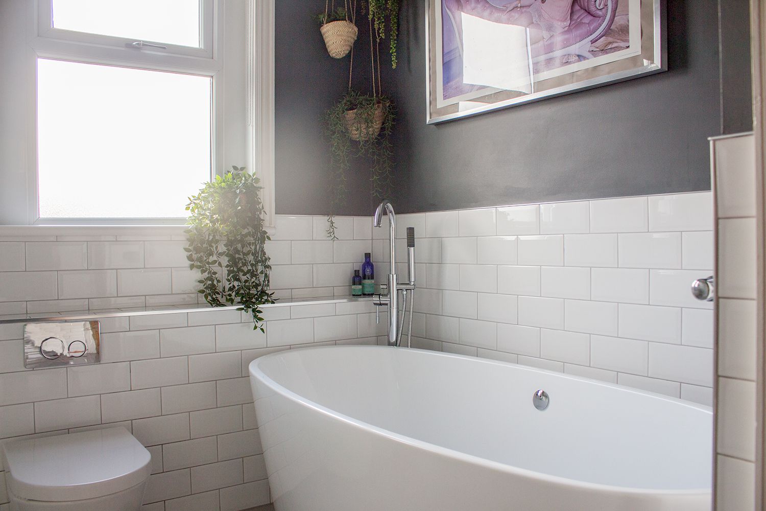 A photo of the bathroom showing the bath, window with new blind, dark walls and metro tiles with hanging planters.