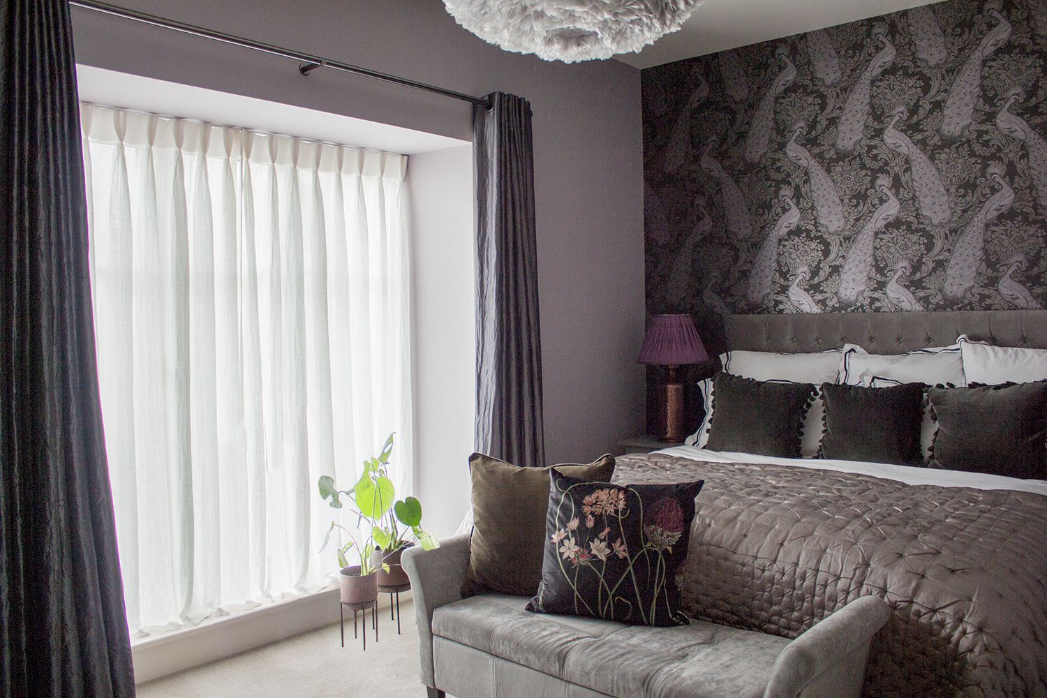 A view of the bed and peacock wallpaper behind. The windows have the original black curtains and sheer curtains added, and some plants in planters.