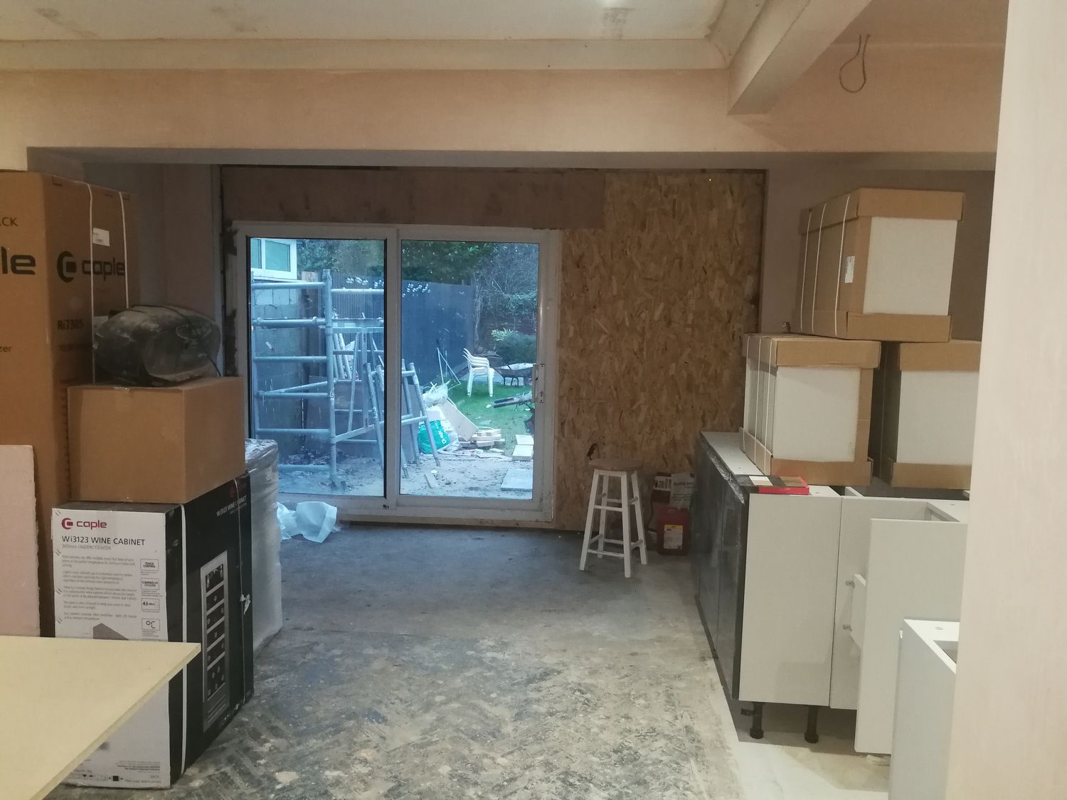 A photo of the same angle with bare plaster walls, some of the kitchen units stacked up and temporary uPVC sliding doors.