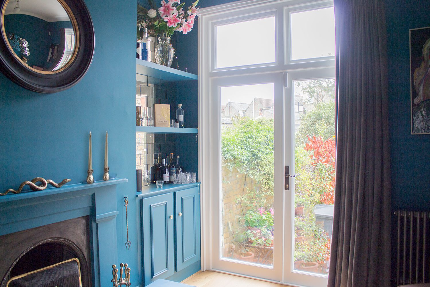 A photo of the teal painted room with a black round mirror over the fireplace and brass accessories.