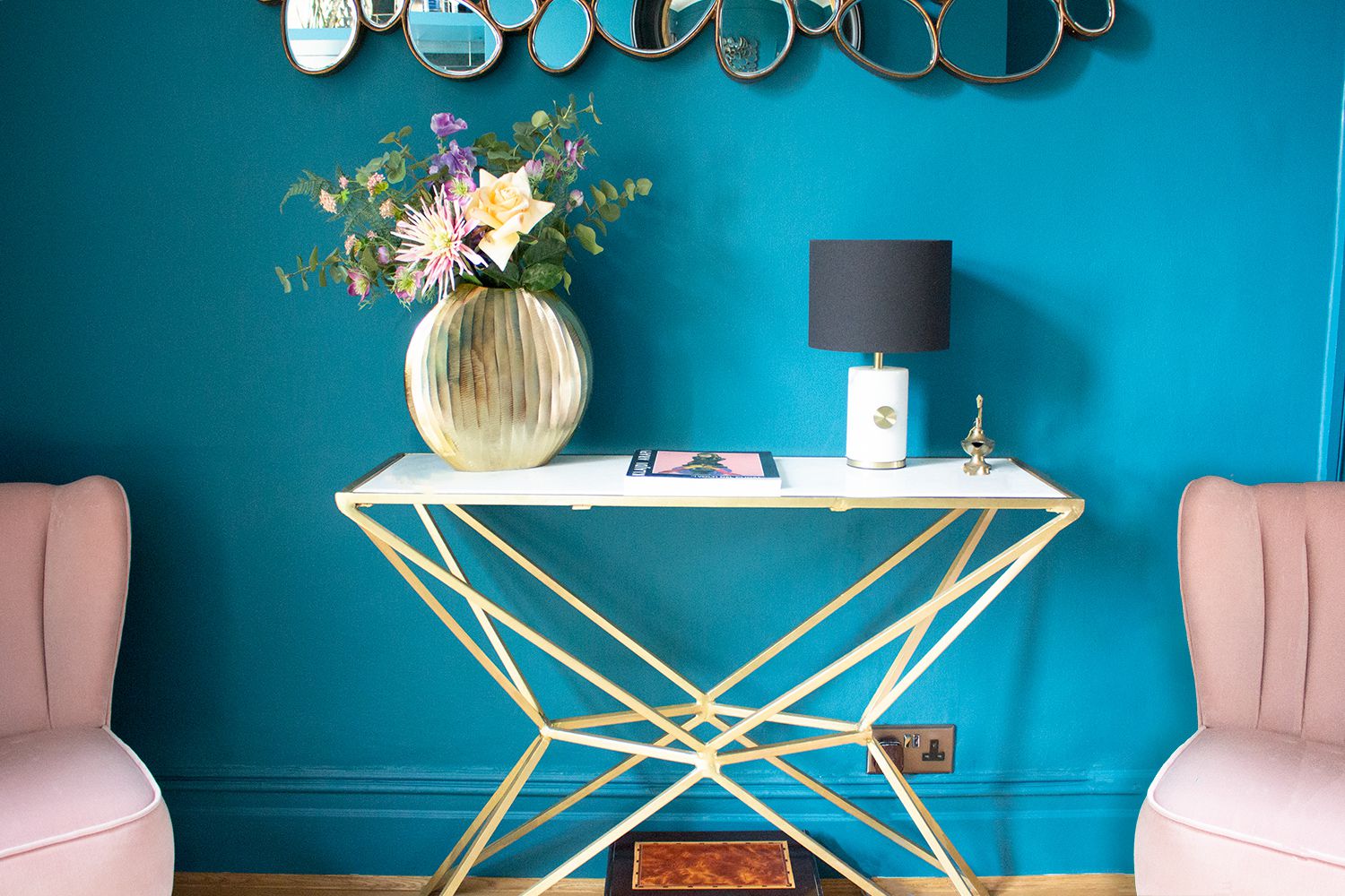 A photo of the teal room with a brass and marble console table and pink velvet cocktail chairs.