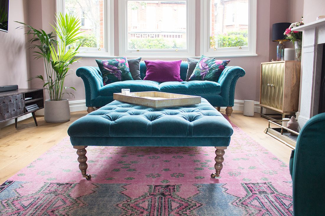 A photo of a teal velvet sofa with matching footstool in a living room with pink walls.