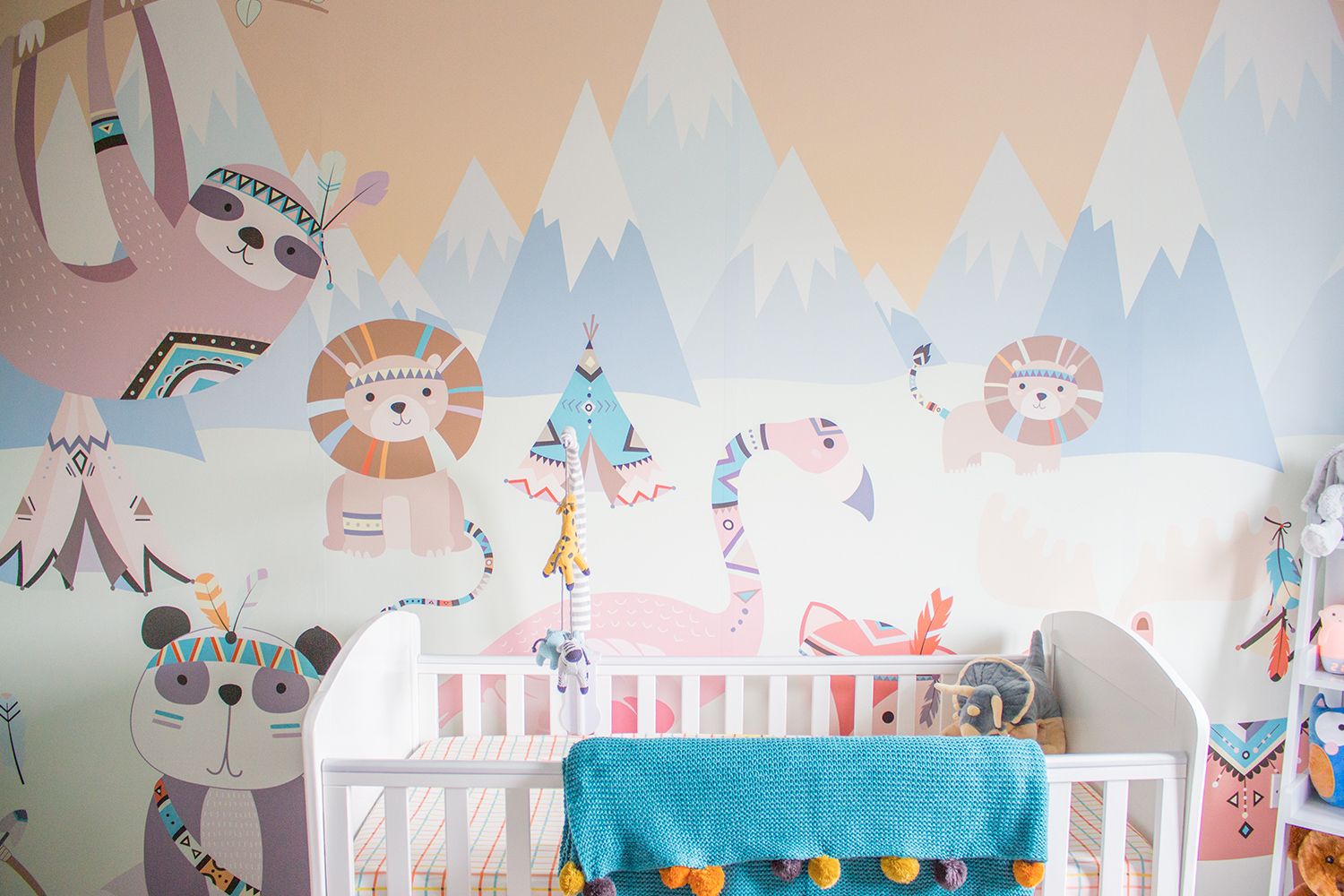 A view of the animal themed wallpaper mural with the white cot in front and colourful blanket draped over the side.