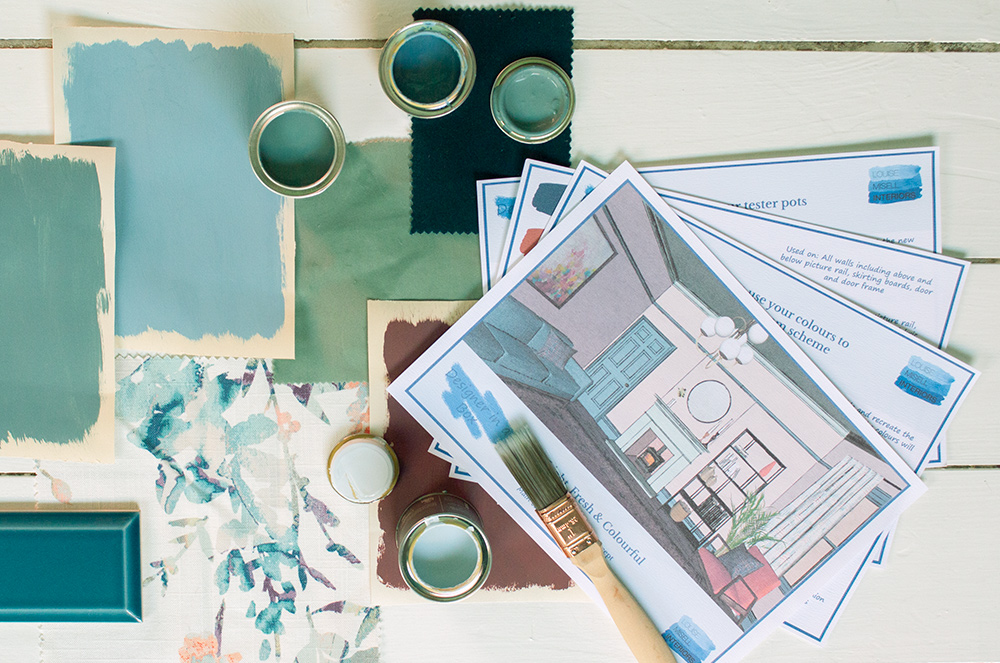 A photo of an example room colour scheme with some open paint pots, a paint brush and some fabrics artfully placed around it.