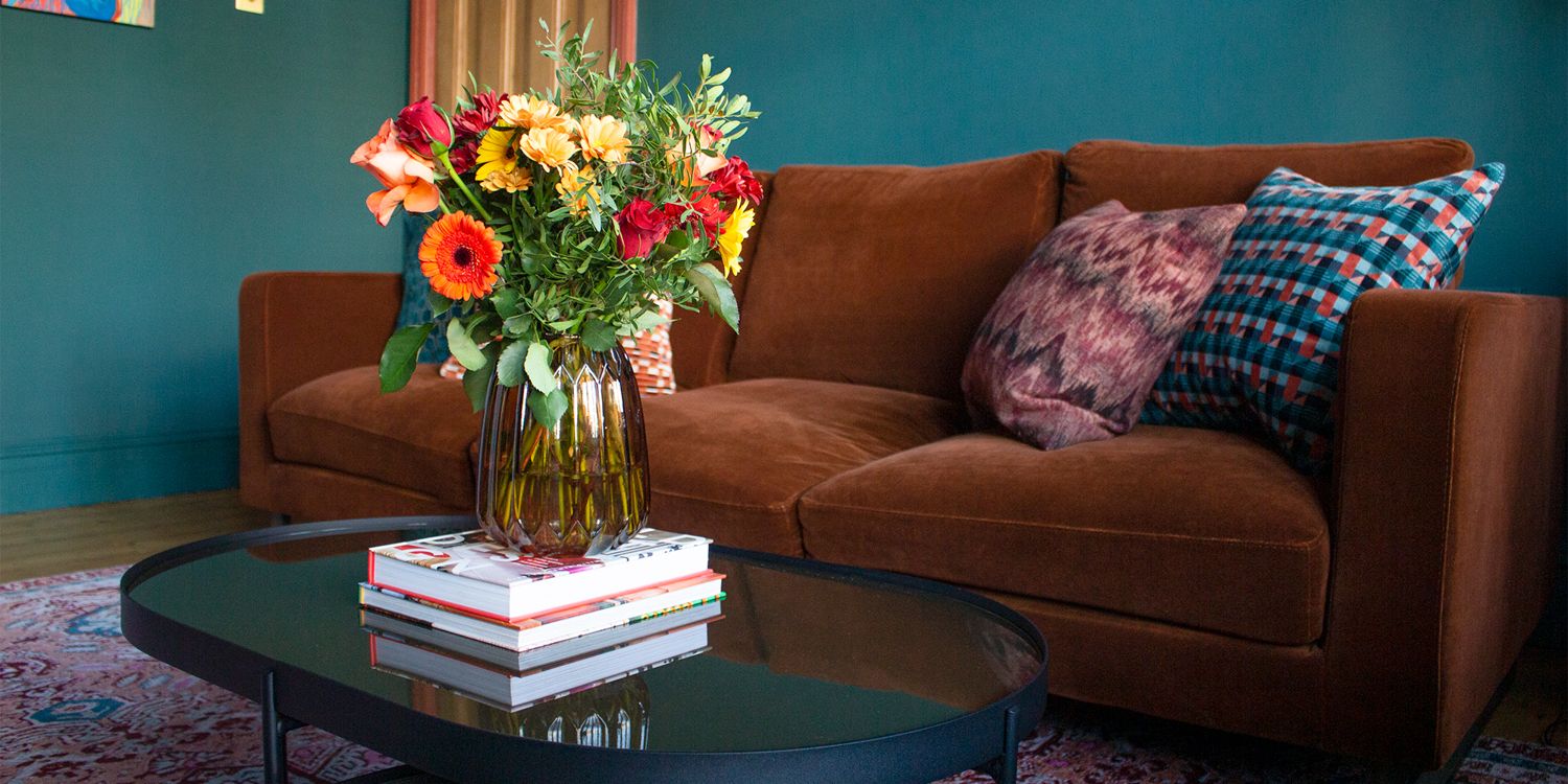 A photo of a cocoa velvet sofa in a room with dark green walls, with a coffee table with flowers on it in front.