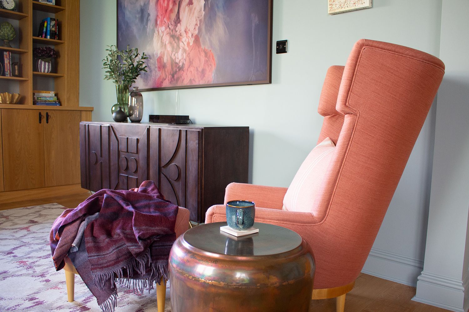 A photo of the study, showing a light red wing chair with matching footstool by a dark wood sideboard.r and