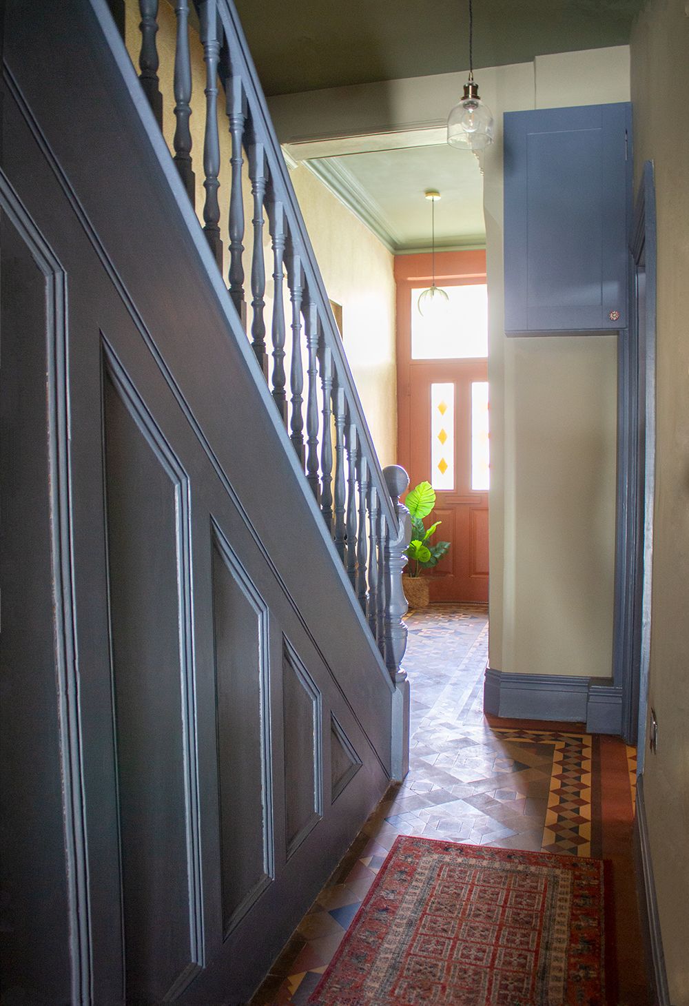 A photo taken looking past the stairs towards the front door. The stairs and paneling are painted blue and there is a red patterned rug on the original quarry tiles.