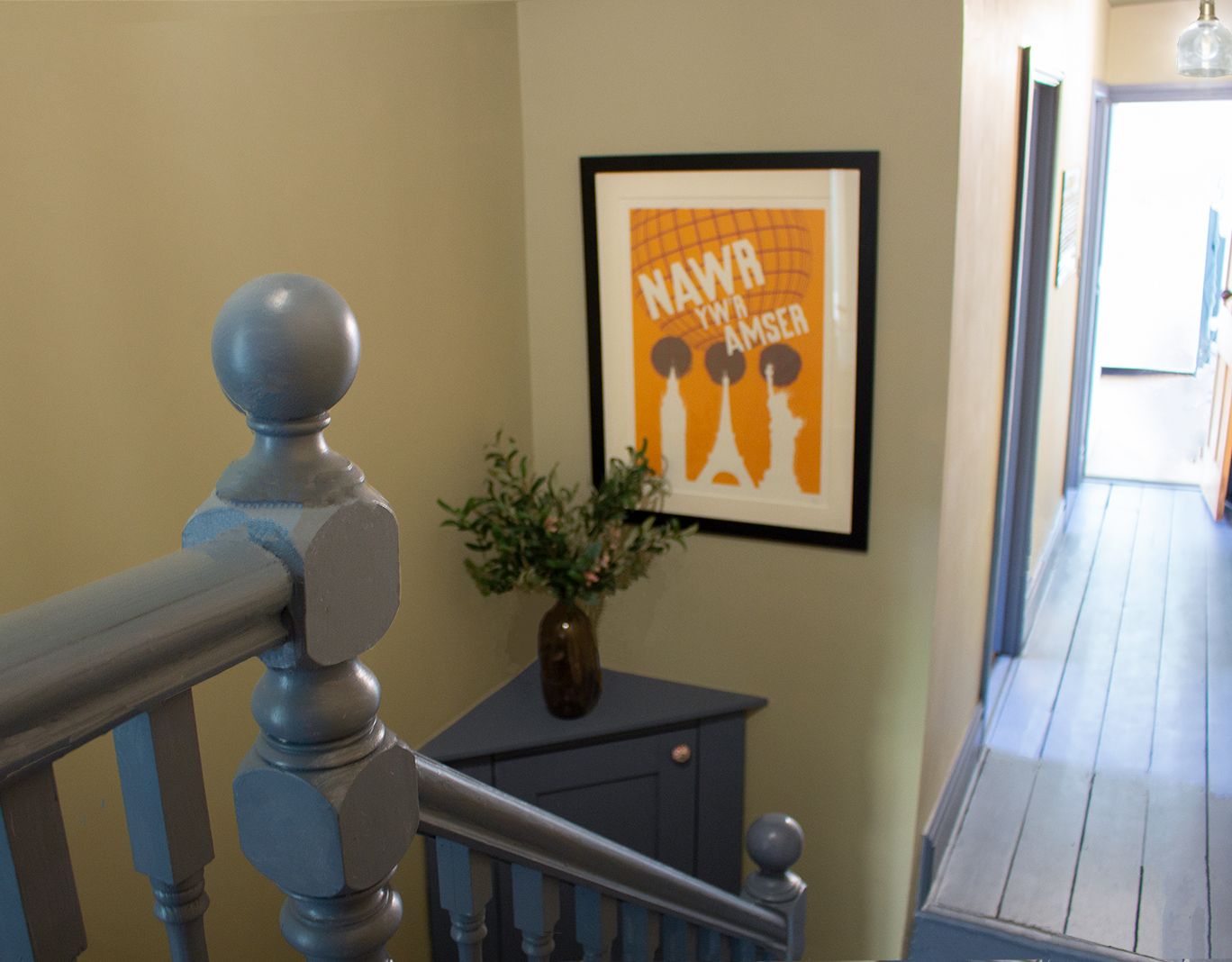 A photo of the landing with blue painted bannisters, skirting boards and built in cupboard. There is a poster on the wall  which says 'now is the time' in Welsh.