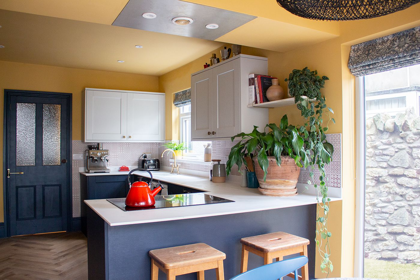 A photo showing the kitchen, with navy blue base units and pale grey wall units. There are William Morris fabric blinds at the windows, and the walls are painted a soft yellow.