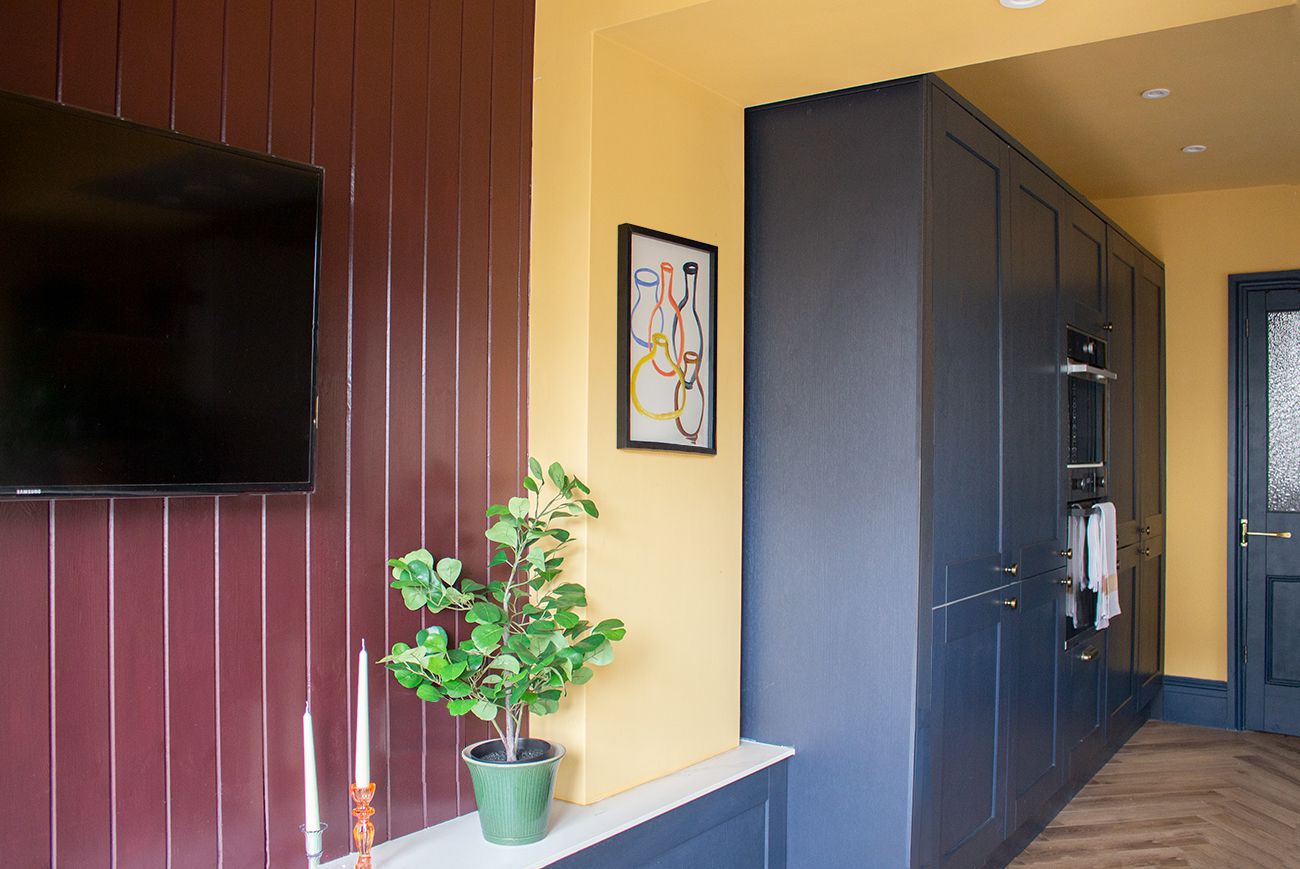 A photo showing the large bank of navy kitchen units and the aubergine painted tongue and groove paneling behind the TV.