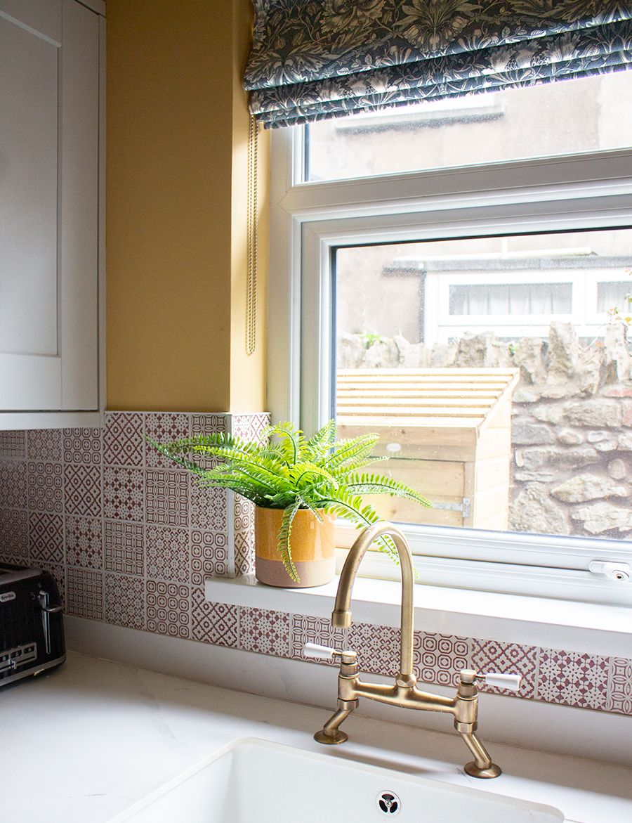 A photo of the sink area with white marble worktops, a red patterned tile on the splash back, and a traditional brass tap.