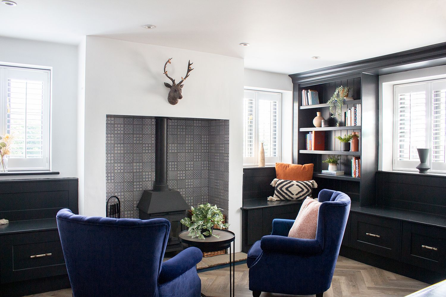 A photo of the snug, with built in shelving and window seats, and blue velvet chairs by the fire.