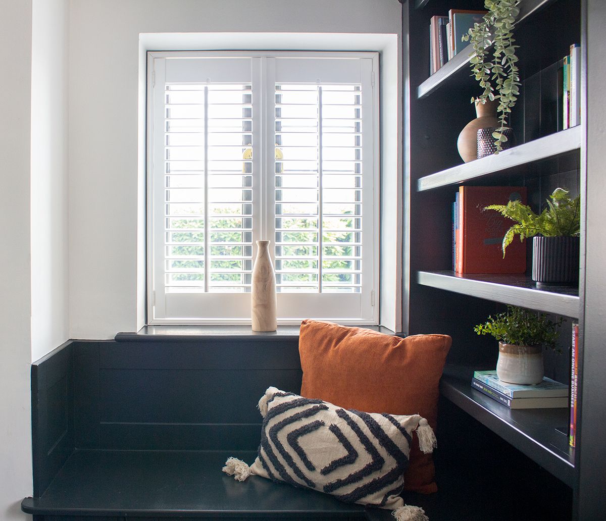 A close up of one of the window seats in the snug, with white shutters at the window and cushions on the seat.