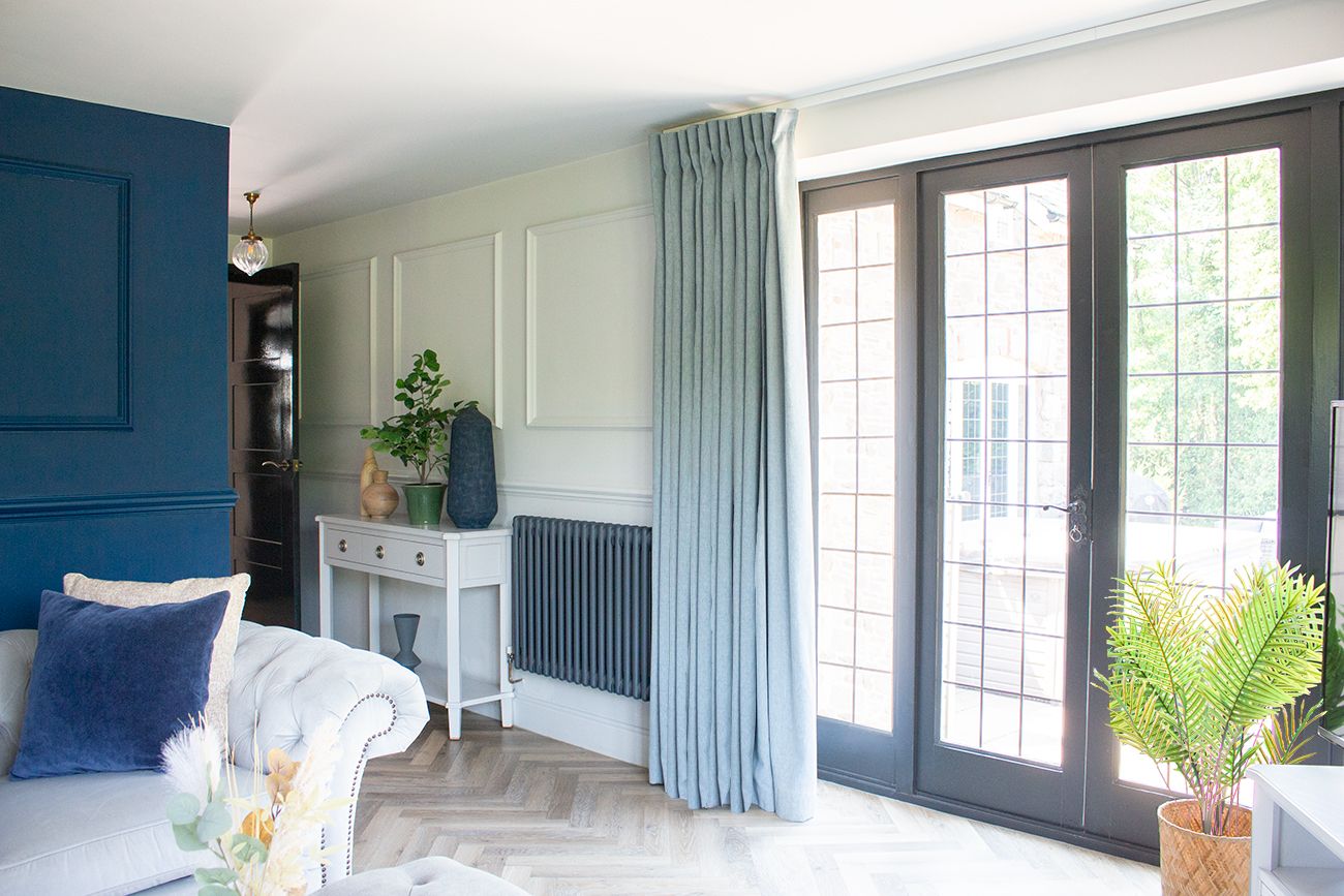 A view of the living room looking back towards the hallway. There are pale blue curtains at the French doors and a console table against a paneled wall.