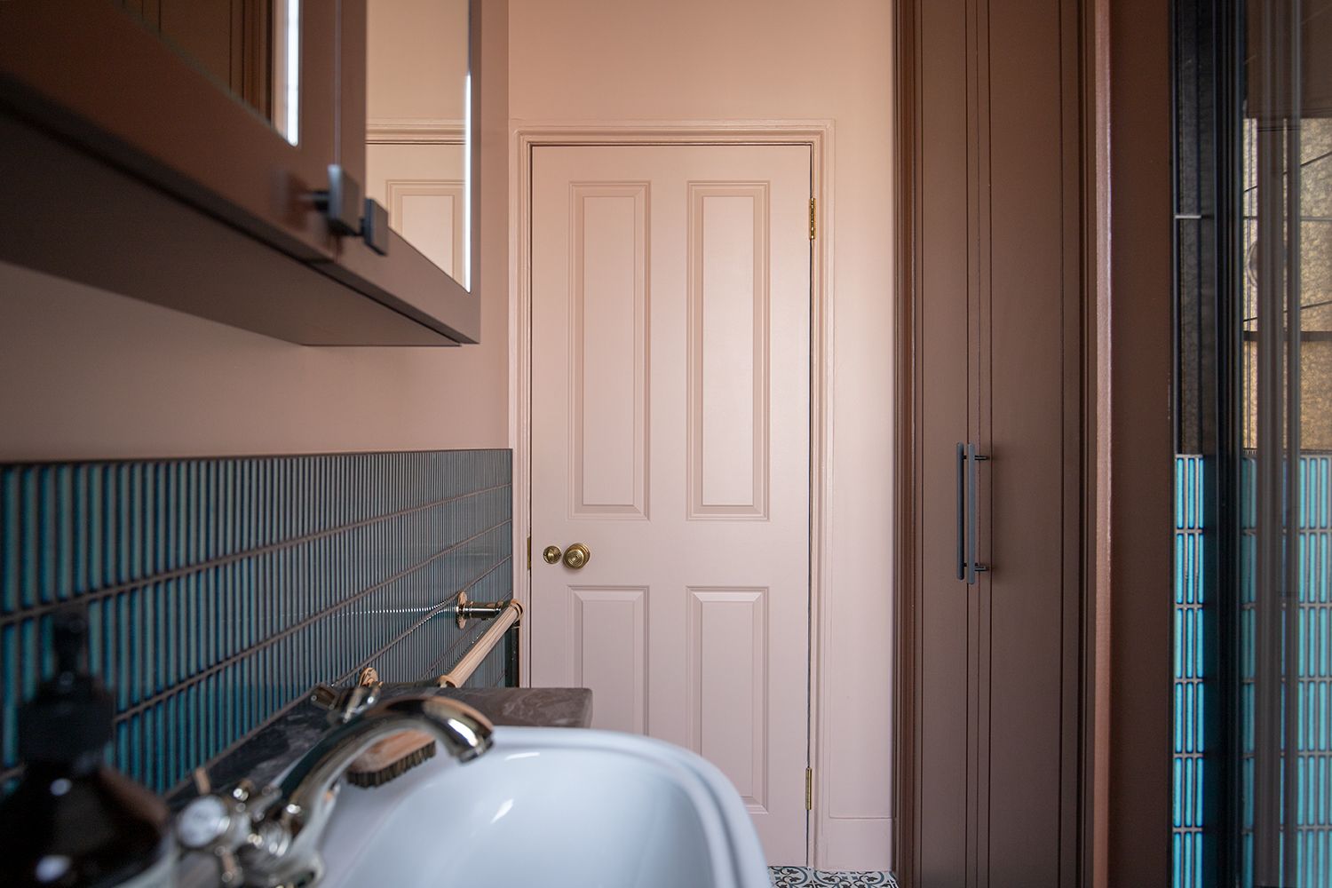 A photo looking along the bathroom cabinets to the back of the door, which is painted in a soft pink to match the walls.